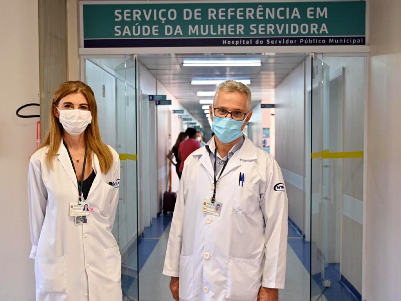 Foto dos médicos Ligia e Ivo na entrada do Serviço de Referência em Saúde da Mulher da HSPM. Eles estão de jaleco branco, máscara e usam crachás. Atrás deles aparece um corredor e algumas pessoas conversando